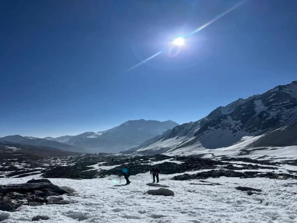 Two trekkers walking across a snowy, rocky mountain moraine under a bright sun and clear blue sky